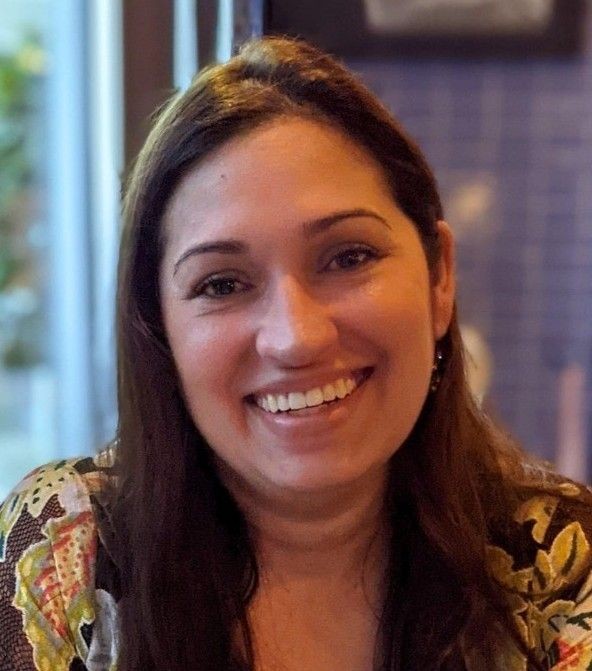 Smiling person with long dark hair and a patterned shirt in a softly lit indoor setting.