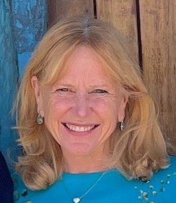 Smiling woman with long blonde hair and earrings, wearing a blue top.